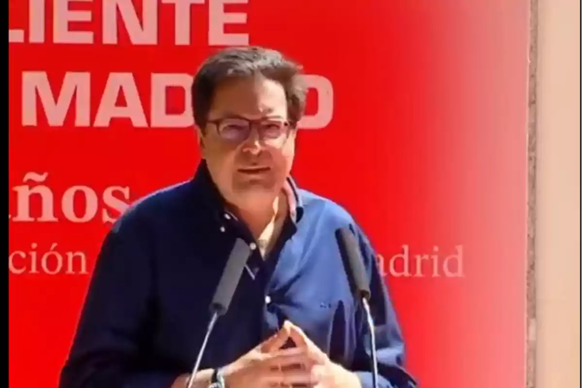 Hombre con gafas y camisa azul hablando frente a dos micrófonos con un fondo rojo y letras blancas Hombre con gafas y camisa azul hablando frente a dos micrófonos con un fondo rojo y letras blancas