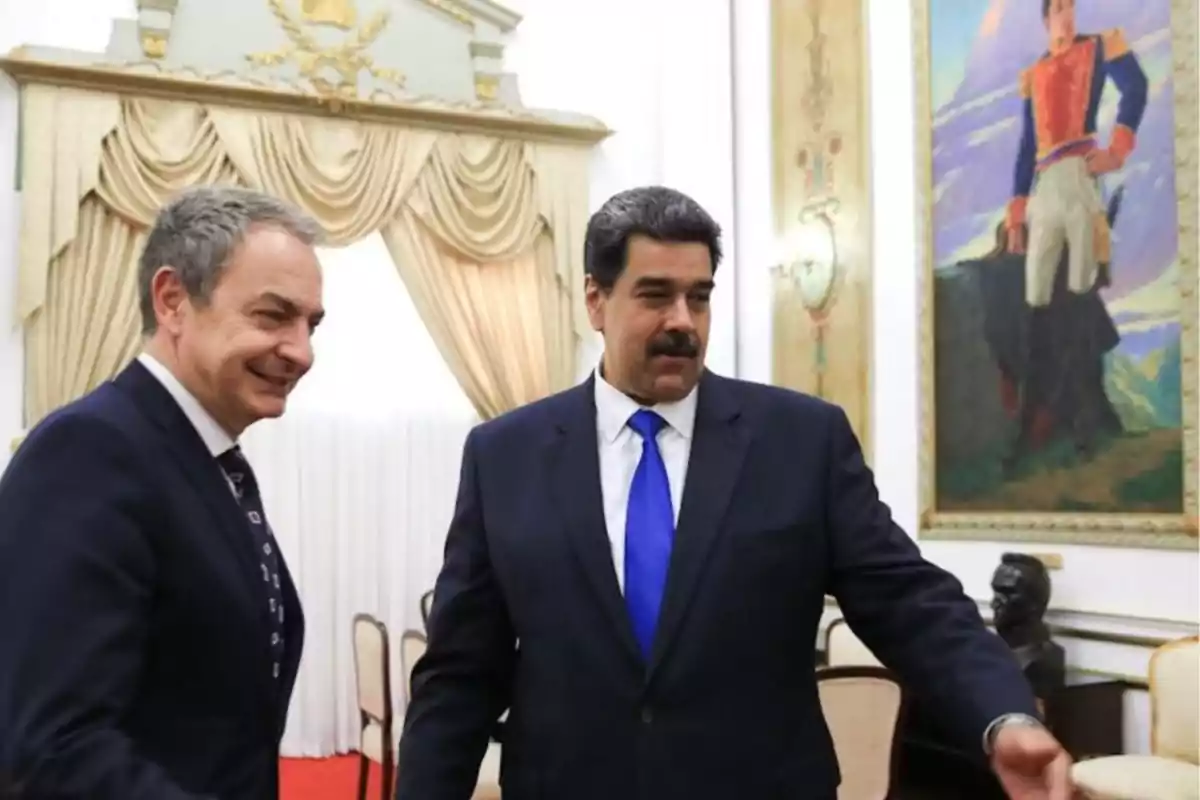 Dos hombres de traje oscuro conversan en una sala decorada con cortinas doradas y un gran cuadro en la pared. Dos hombres de traje oscuro conversan en una sala decorada con cortinas doradas y un gran cuadro en la pared.